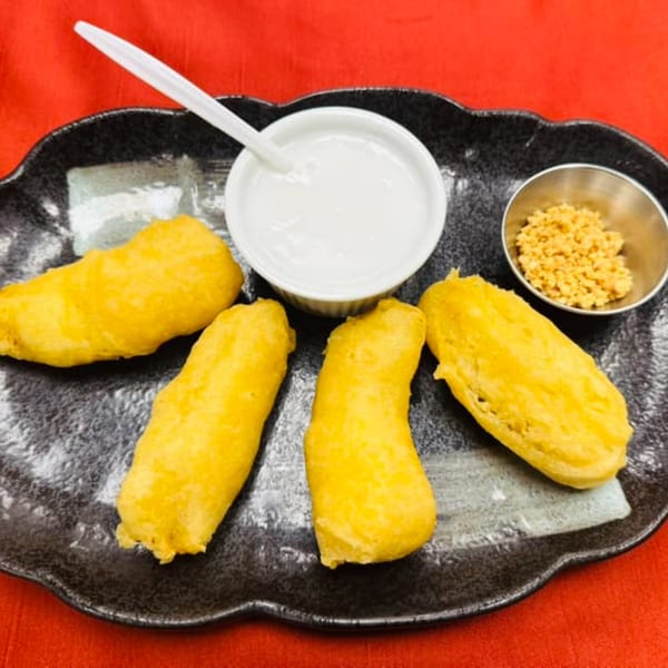 Four golden fried bananas with a side of coconut cream and crushed peanuts on a black plate.