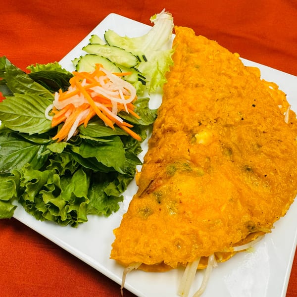 Golden Vietnamese savory crepe (Banh Xeo) with fresh herbs, pickled carrots, and daikon on a white plate.