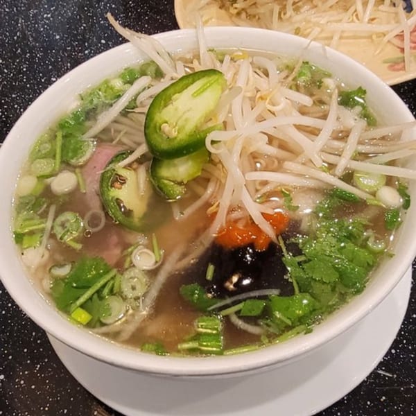 Vietnamese pho with bean sprouts, cilantro, green onions, jalapeños, and sauces in a white bowl.