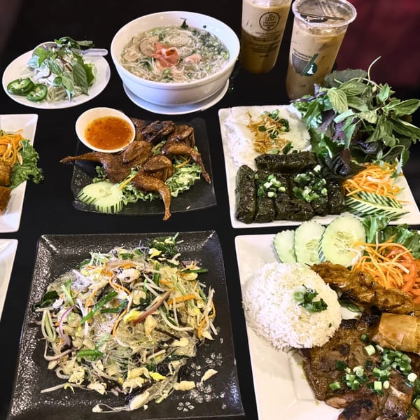 A table spread of Vietnamese dishes including pho, grilled quail, vermicelli, and rice plates with iced coffee.