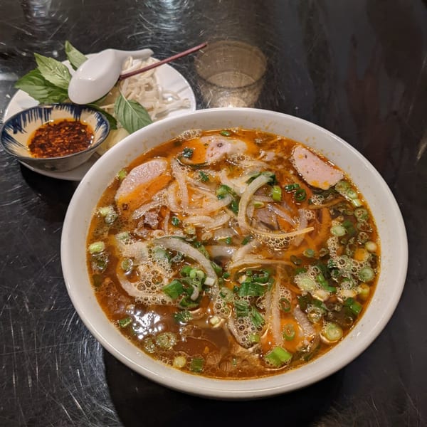 A bowl of spicy Vietnamese pho with meat, noodles, and green onions, with a side of herbs.